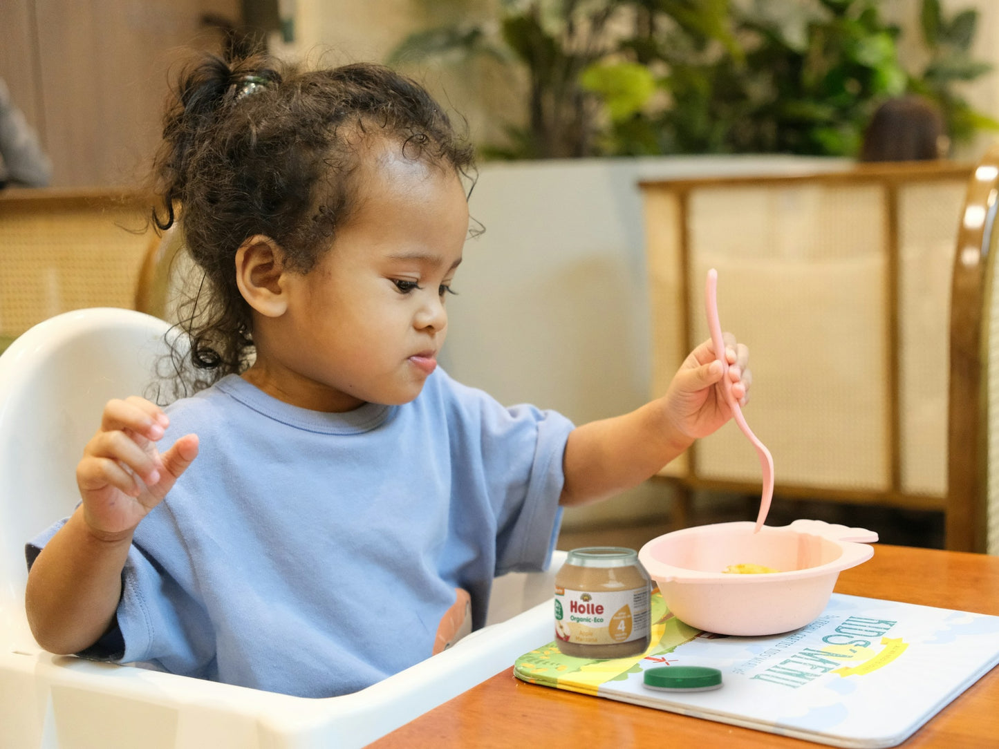 Toddler in a high chair eating from a pink bowl next to an open organic baby food jar